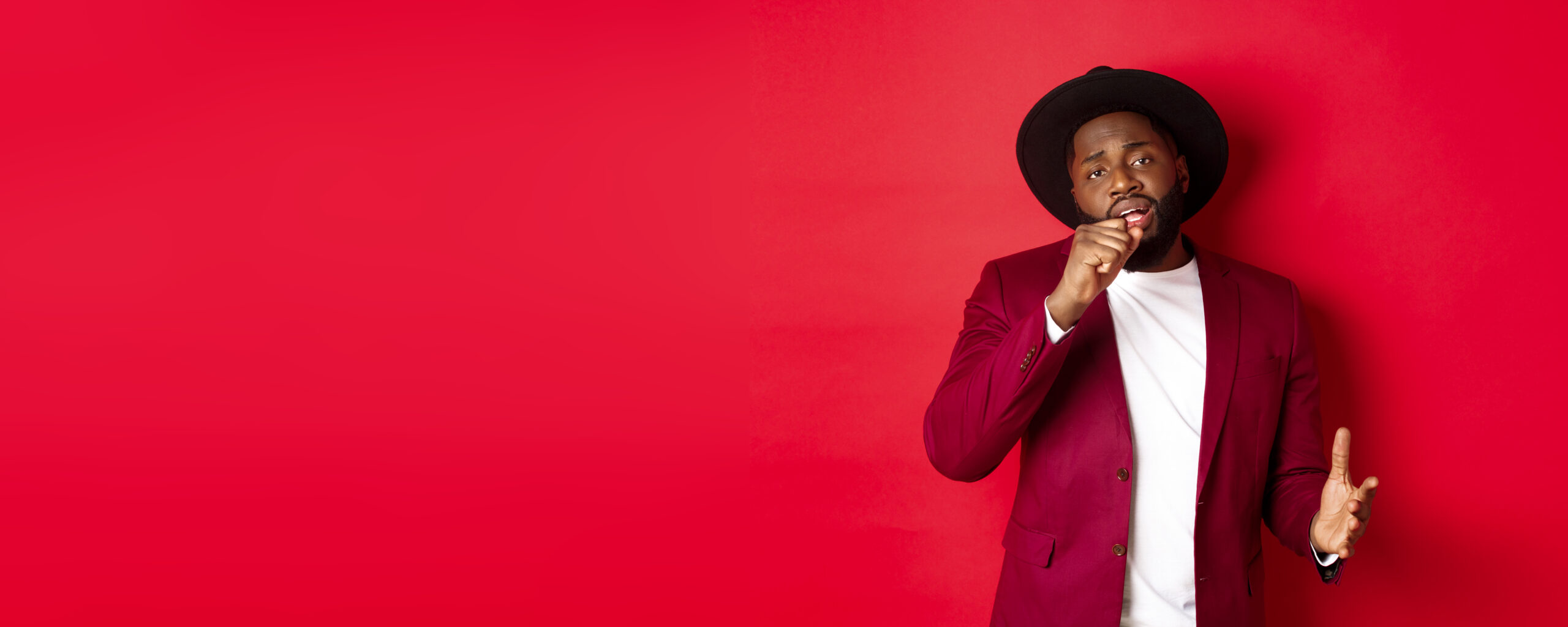 Christmas shopping and people concept. Handsome Black male model in party outfit singing in invisible microphone, looking passionate at camera, red background.